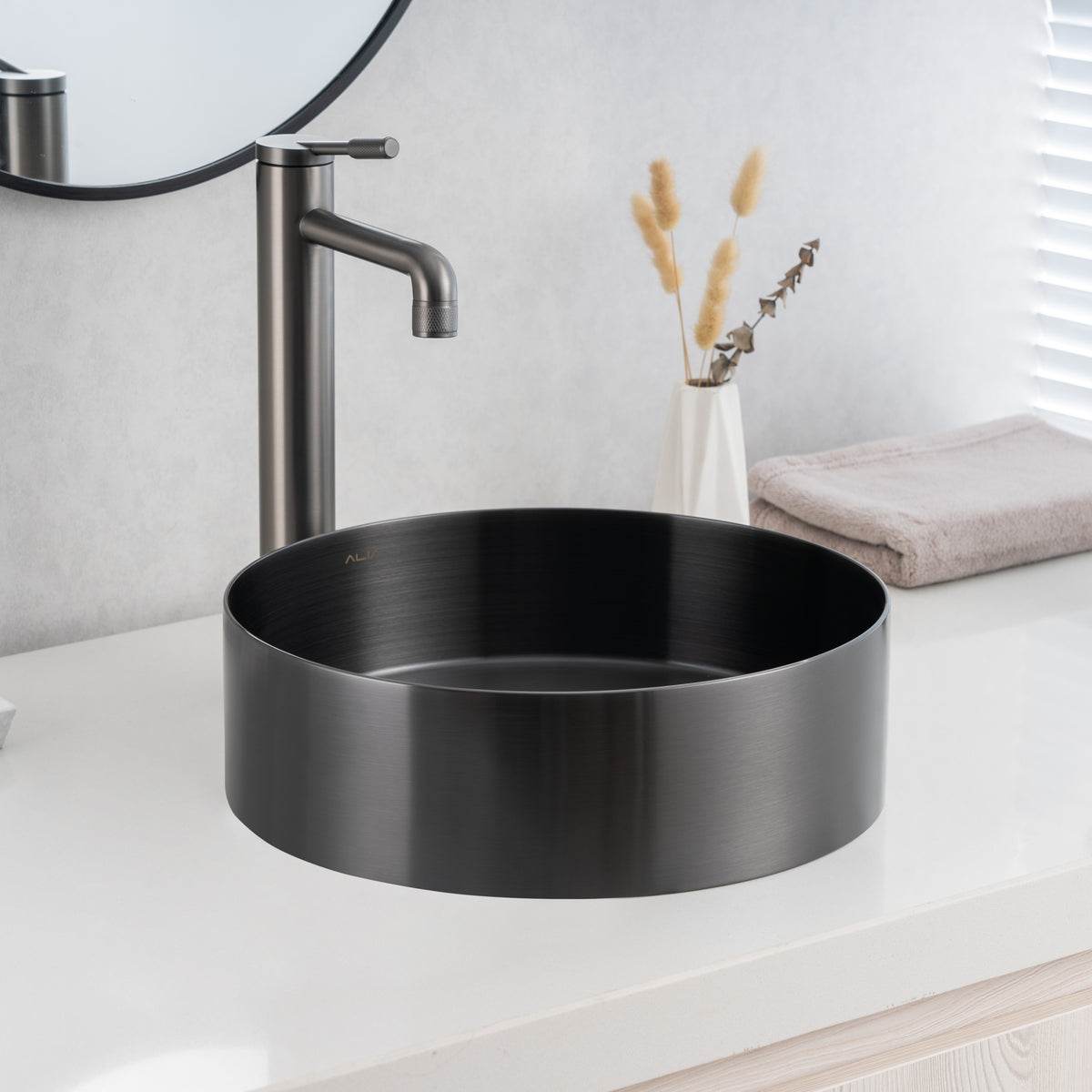 ALIA gunmetal grey round basin offers a durable, easy-to-clean stainless steel finish ideal for designer bathroom sinks. Black round sink on a bathroom counter with a mirror and towel in the background.